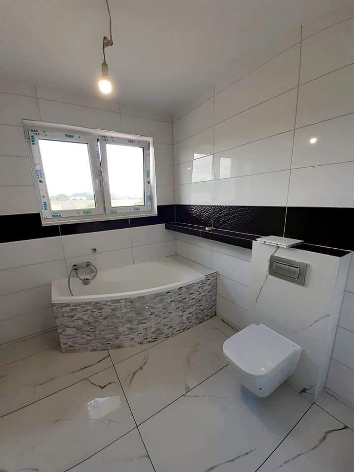 Wykończona łazienka z białymi płytkami imitującymi marmur na podłodze i ścianach, z czarnym pasem płytek dekoracyjnych na ścianie, widoczna wanna obudowana mozaiką, wisząca toaleta i okno...