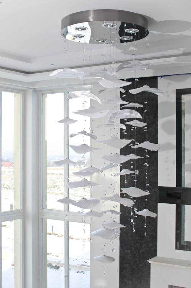 Nowoczesny żyrandol z okrągłą, metalową podstawą i zwisającymi, białymi elementami przypominającymi kapelusze, ozdobiony kryształkami, w jasnym wnętrzu z dużymi oknami.