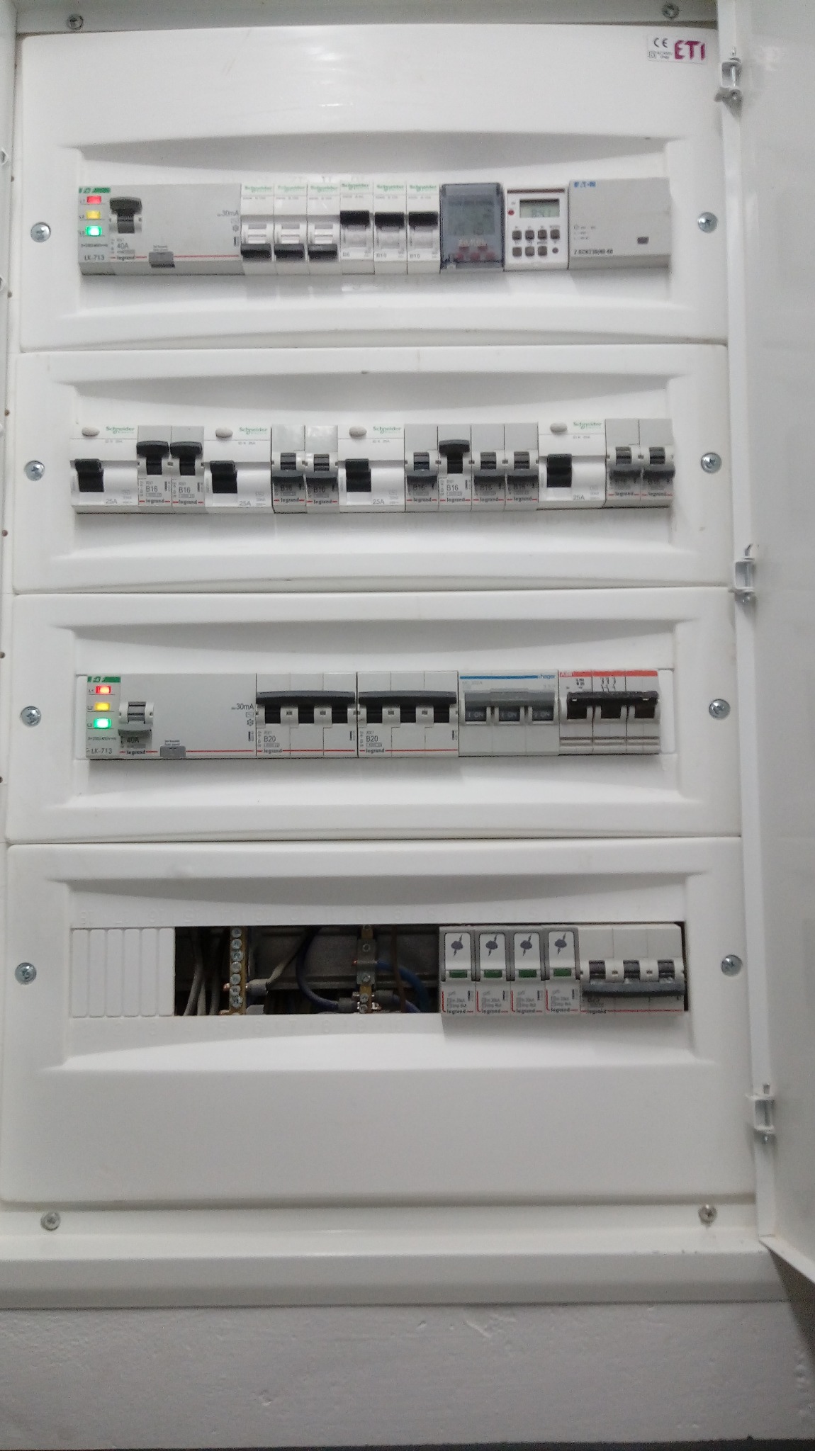 Otwarta skrzynka elektryczna z czterema rzędami zabezpieczeń i różnicówkami marki Schneider Electric i Legrand, widoczne przewody w dolnej części.