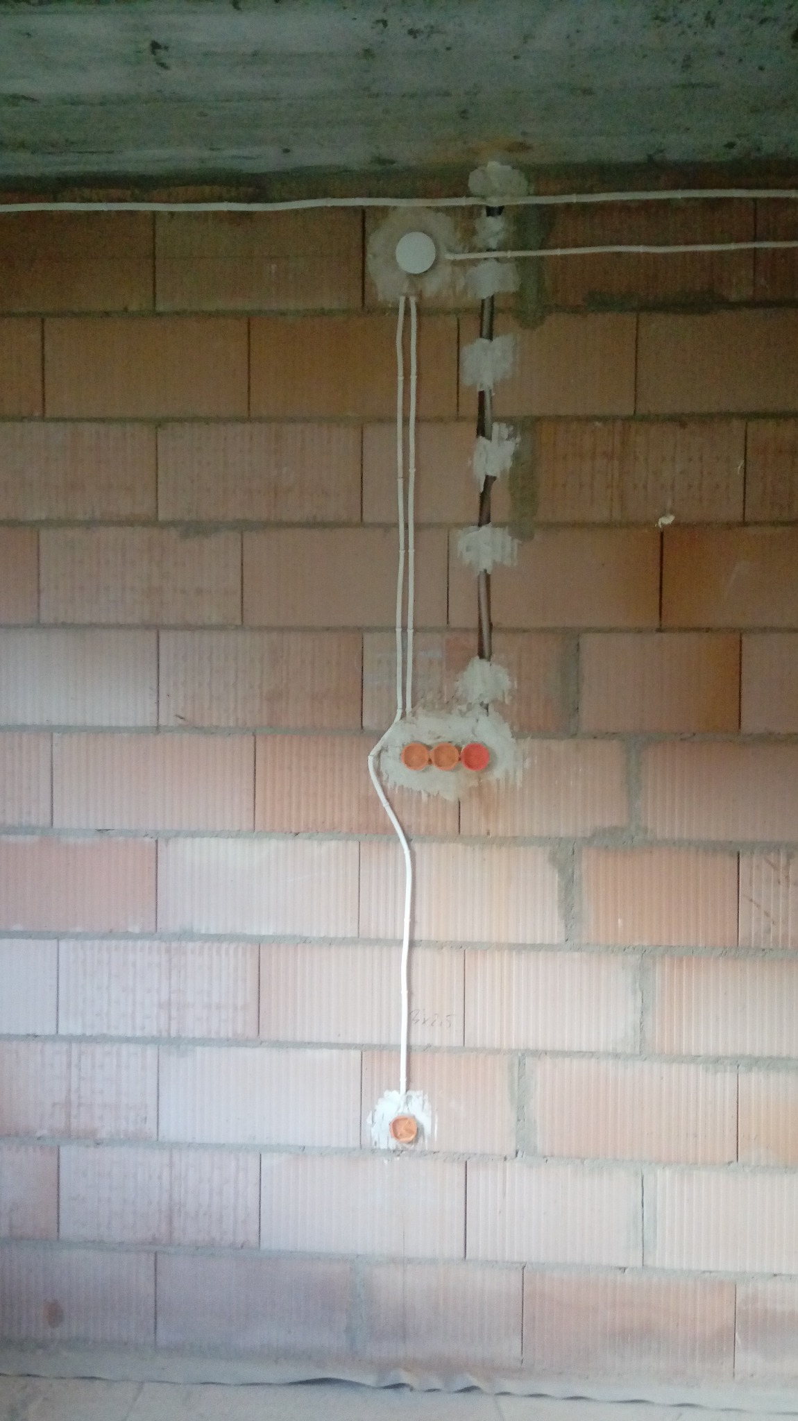 Instalacja elektryczna w trakcie montażu w surowym murze z cegły, widoczne przewody w peszlu, puszki instalacyjne z pomarańczowymi wkładkami i białe kable.
