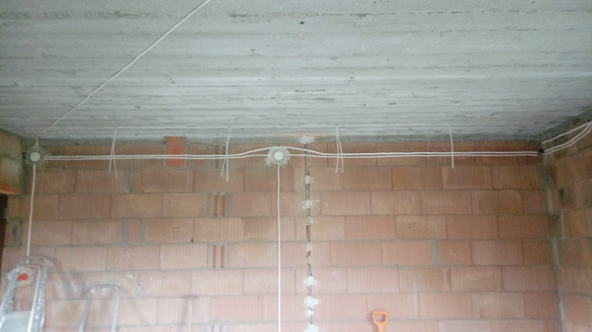 Surowy stan: Instalacja elektryczna w trakcie montażu w pomieszczeniu z ceglanymi ścianami i betonowym sufitem, widoczne puszki instalacyjne i przewody, drabina w tle.