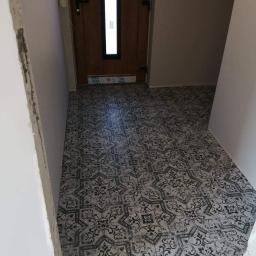 Waldemar Malina - Korytarz z nowo ułożoną, wzorzystą podłogą ceramiczną w odcieniach szarości i czerni, widoczne niedokończone prace wykończeniowe ścian i okleina ochronna na ościeżnicy drzwi.