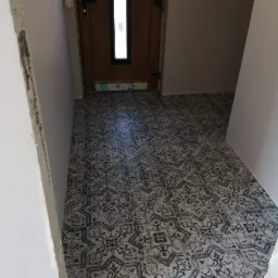 Korytarz z nowo ułożoną, wzorzystą podłogą ceramiczną w odcieniach szarości i czerni, widoczne niedokończone prace wykończeniowe ścian i okleina ochronna na ościeżnicy drzwi.