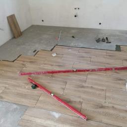Waldemar Malina - Układanie paneli podłogowych imitujących drewno w trakcie prac, widoczne poziomice, krzyżyki dystansowe i fragmenty nieukończonej posadzki.