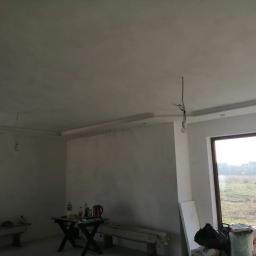 Waldemar Malina - Pokój w trakcie prac wykończeniowych: świeżo położona gładź na ścianach i suficie, widoczne przewody elektryczne, okno z widokiem na zewnątrz, stolik z narzędziami i napojami oraz materiały...