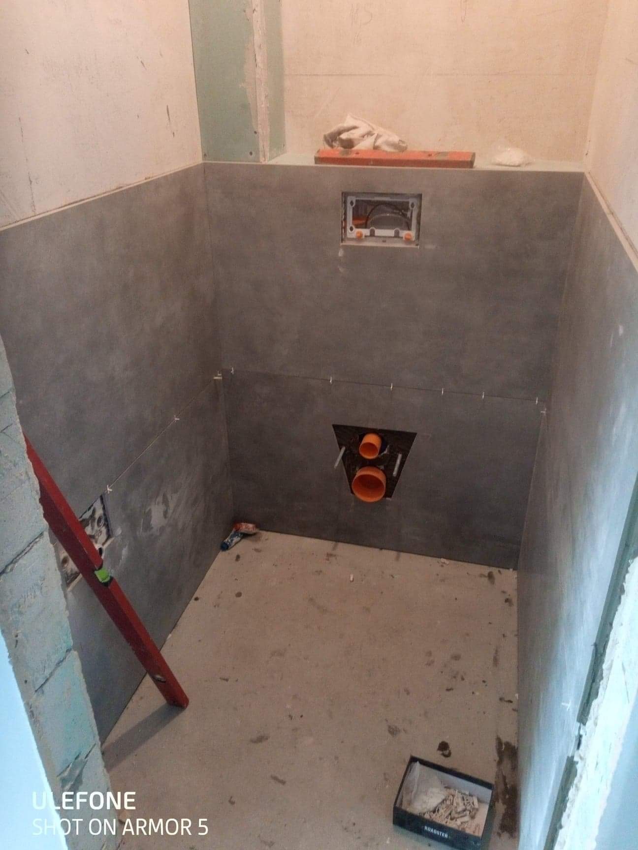 Remont łazienki w trakcie: Szare płytki ścienne częściowo ułożone, widoczne otwory na instalacje sanitarne, poziomica oparta o ścianę, pudełko z resztkami płytek na podłodze.