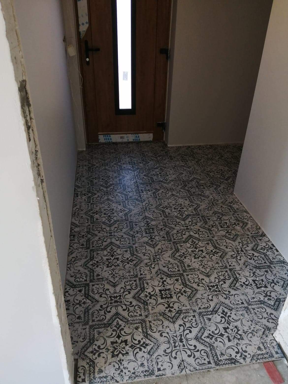 Korytarz z nowo ułożoną, wzorzystą podłogą ceramiczną w odcieniach szarości i czerni, widoczne niedokończone prace wykończeniowe ścian i okleina ochronna na ościeżnicy drzwi.
