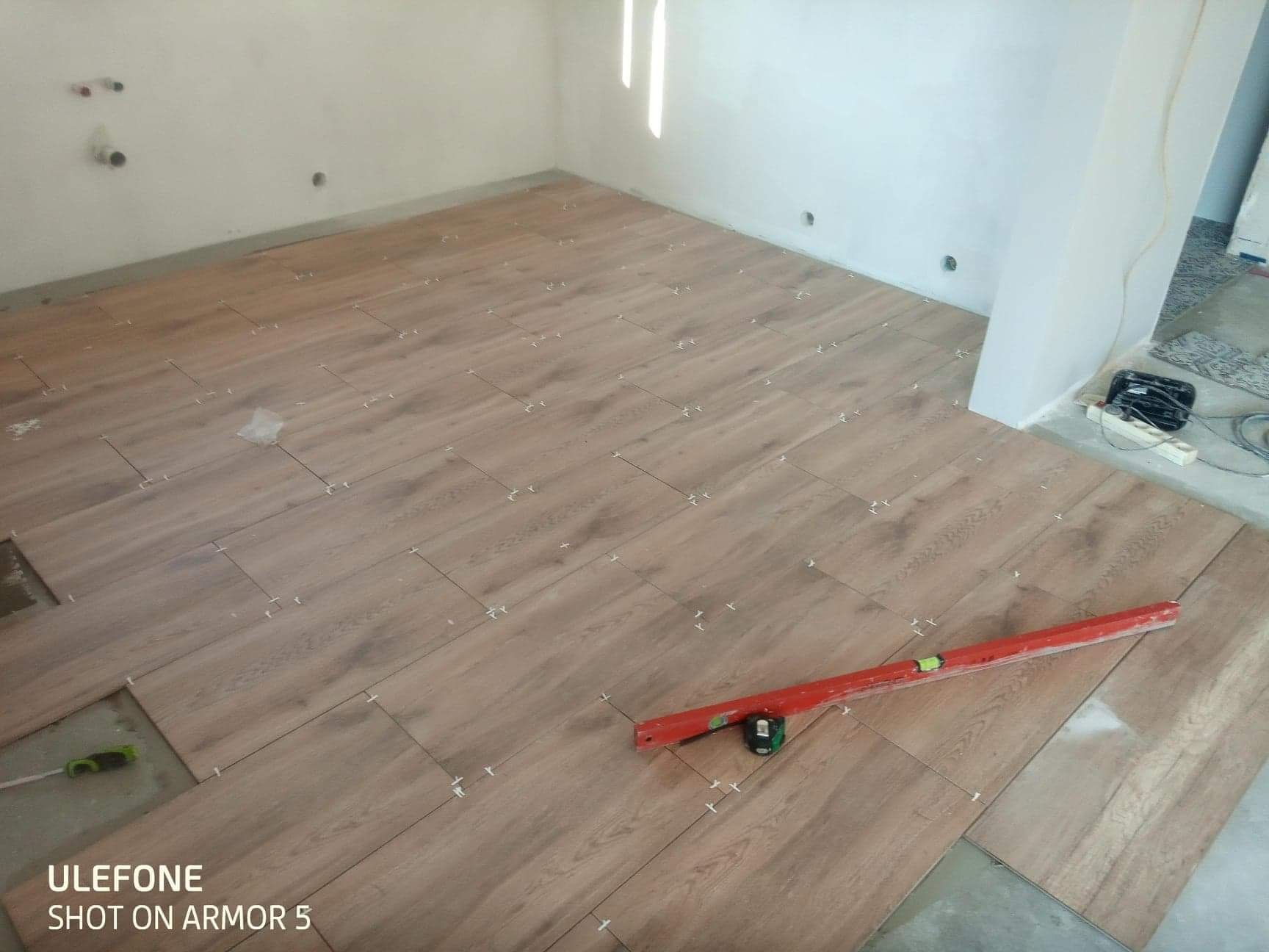 Układanie paneli podłogowych imitujących drewno, z użyciem krzyżyków dystansowych i poziomicy, widoczne niedokończone fragmenty oraz gniazdka elektryczne w ścianie.
