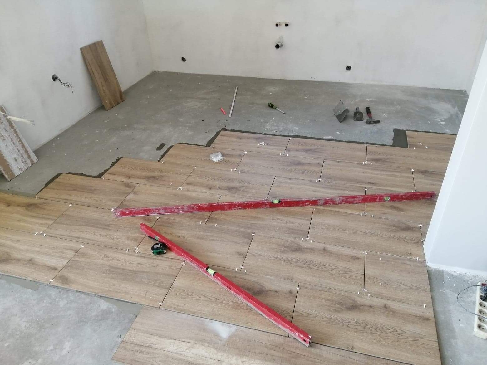 Układanie paneli podłogowych imitujących drewno w trakcie prac, widoczne poziomice, krzyżyki dystansowe i fragmenty nieukończonej posadzki.