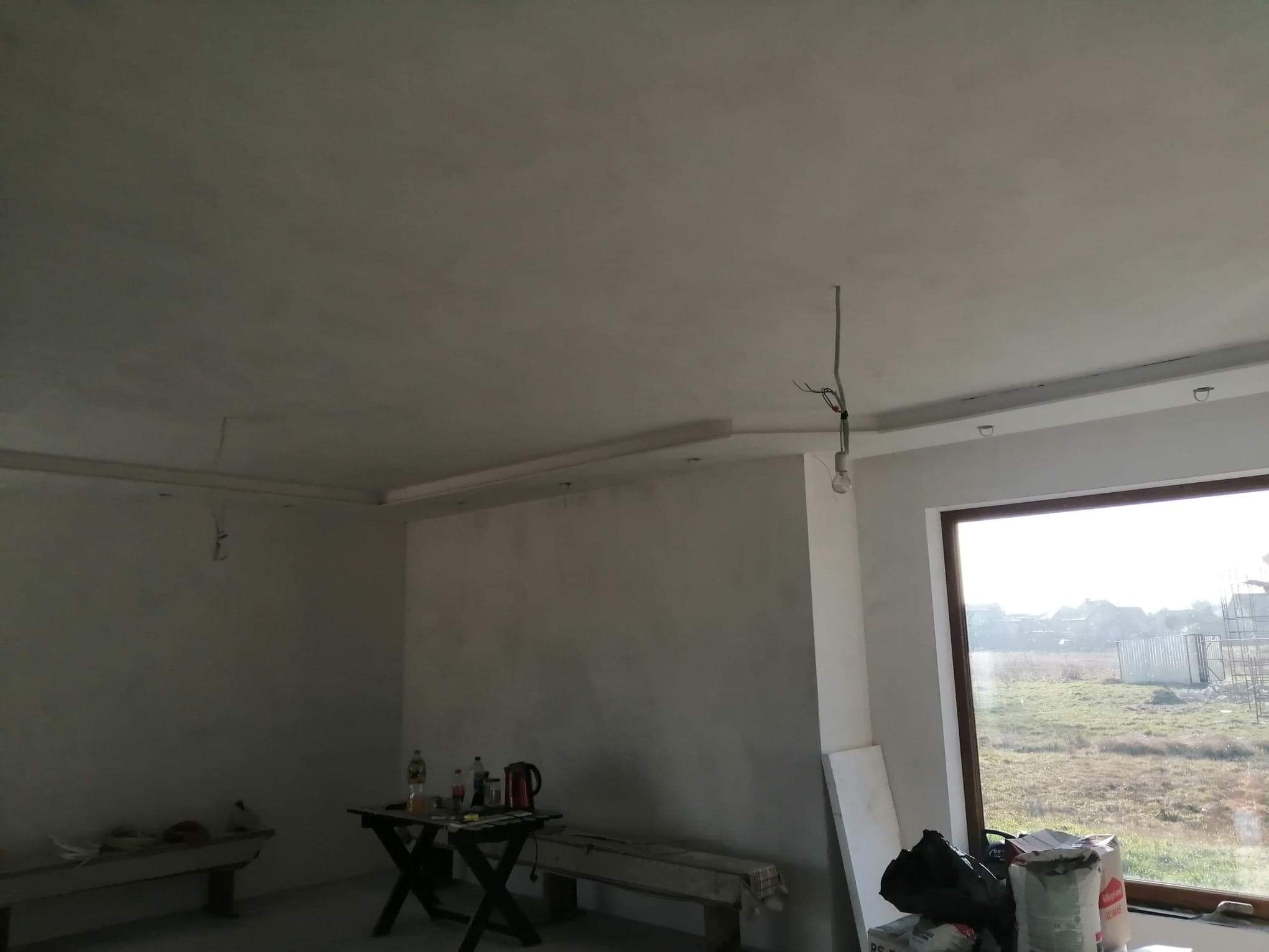 Pokój w trakcie prac wykończeniowych: świeżo położona gładź na ścianach i suficie, widoczne przewody elektryczne, okno z widokiem na zewnątrz, stolik z narzędziami i napojami oraz materiały...