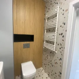 Nowoczesna łazienka z wiszącą toaletą, grzejnikiem drabinkowym na ścianie w stylu lastryko oraz zabudową meblową z drewna.