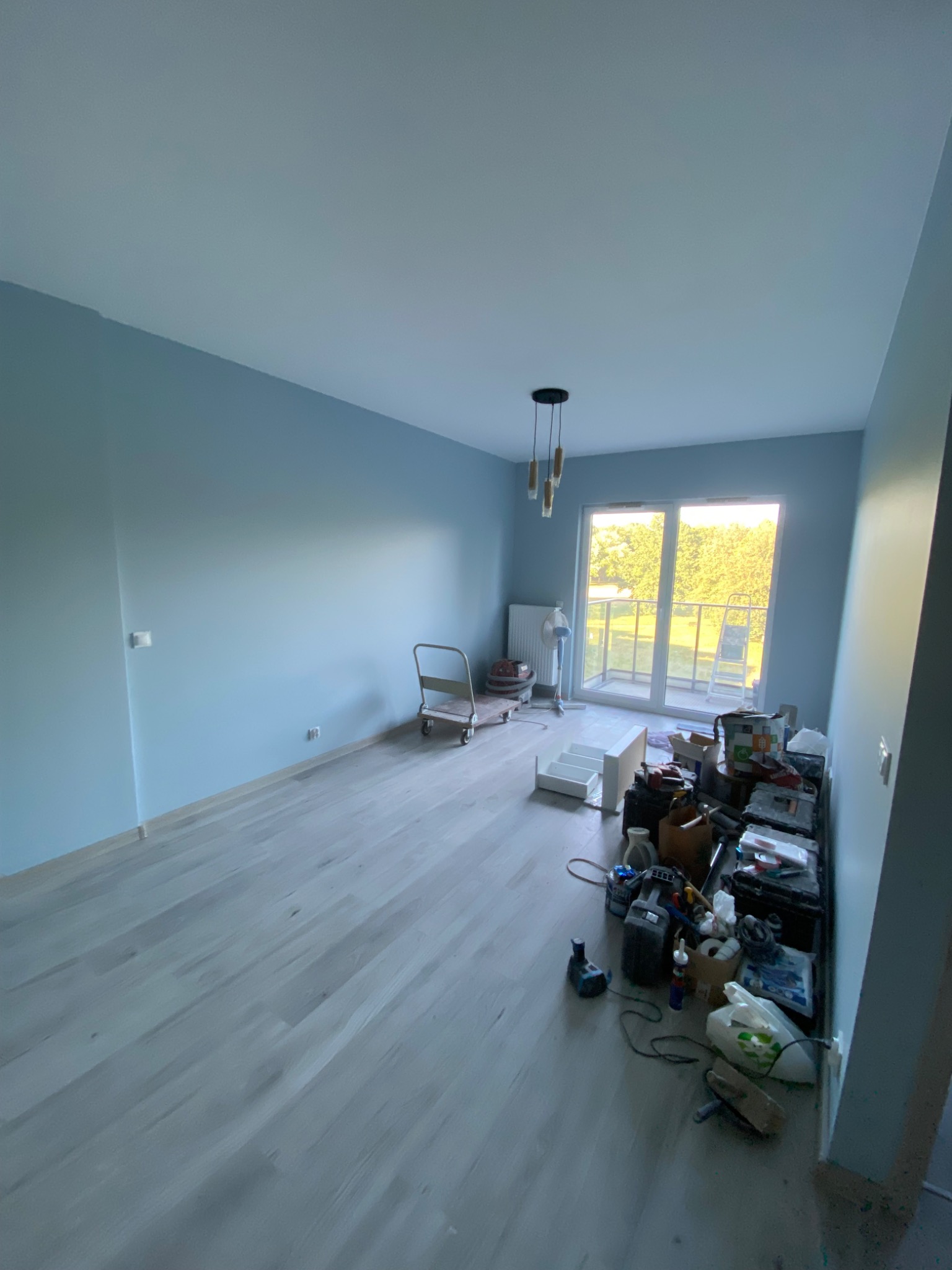 Pokój po malowaniu ścian na jasny niebieski kolor, widoczne narzędzia i akcesoria malarskie oraz świeżo położona jasna podłoga laminowana, balkon z widokiem na zieleń w tle.