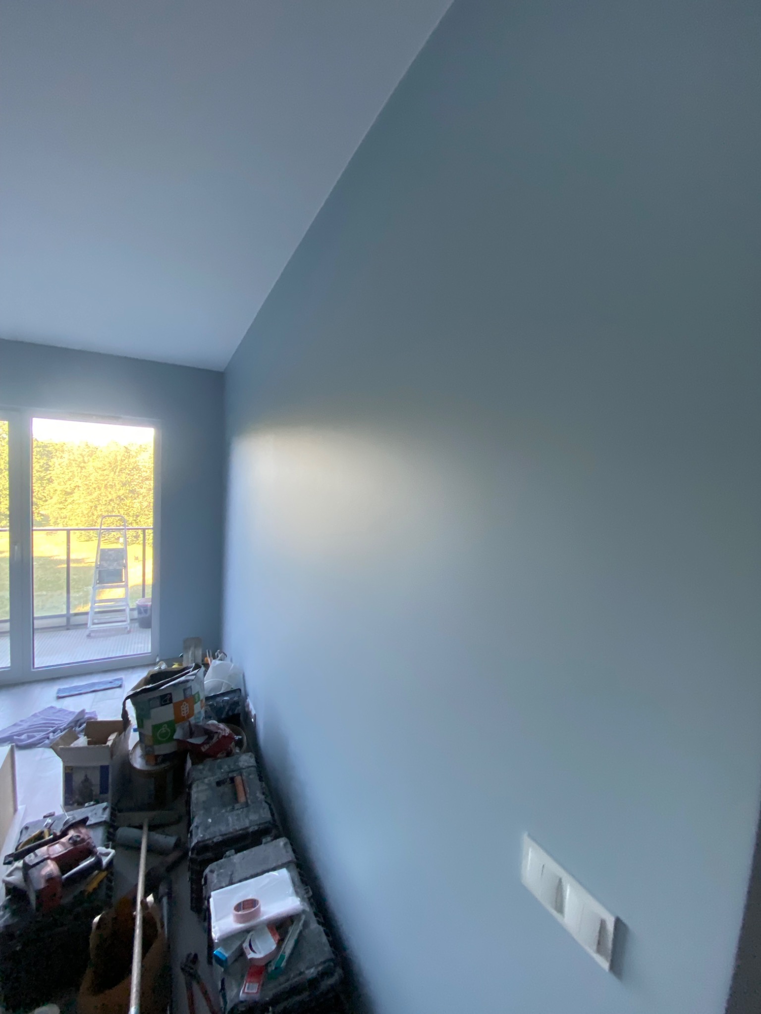 Świeżo pomalowane na błękitno wnętrze z widocznymi narzędziami malarskimi i drabiną na balkonie w tle, sugerujące prace wykończeniowe.