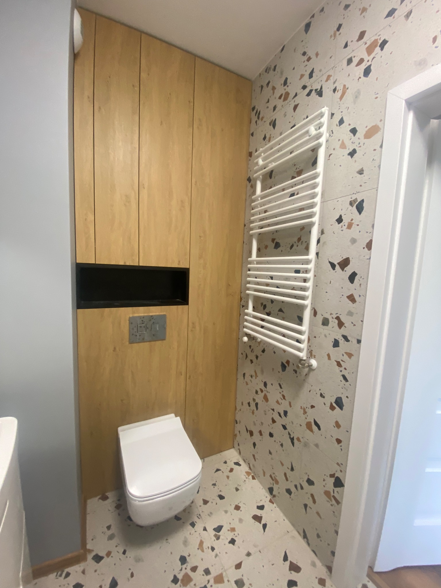 Nowoczesna łazienka z wiszącą toaletą, grzejnikiem drabinkowym na ścianie w stylu lastryko oraz zabudową meblową z drewna.