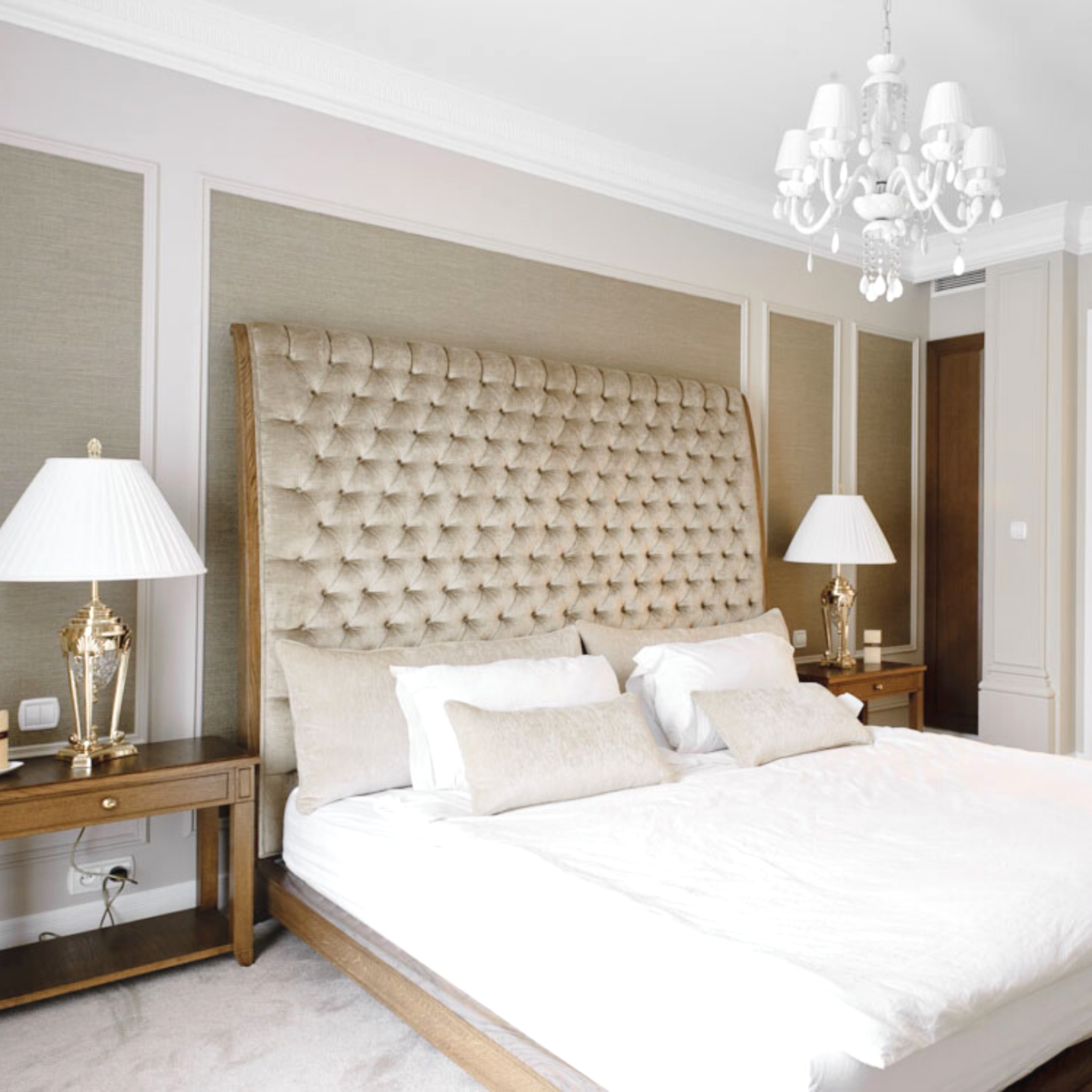 Elegancka sypialnia w jasnych barwach z dużym tapicerowanym zagłówkiem, drewnianymi szafkami nocnymi i kryształowym żyrandolem.