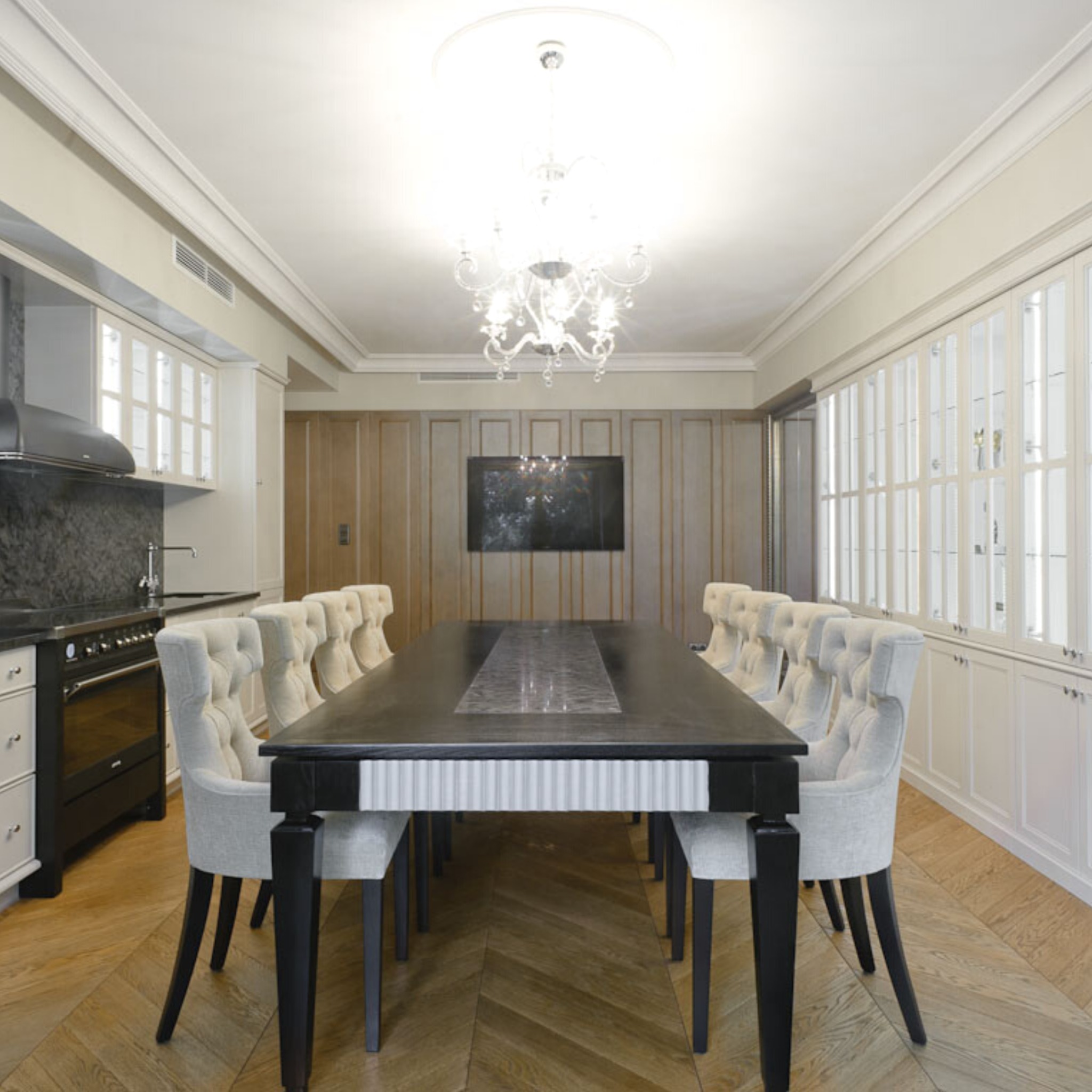 Elegancka jadalnia z długim, ciemnym stołem otoczonym jasnymi, tapicerowanymi krzesłami, parkiet w jodełkę, kuchnia w tle z czarnym blatem i białymi szafkami, ozdobny żyrandol.