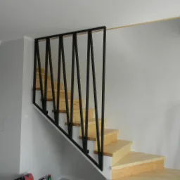 Drewniane schody z czarną, geometryczną balustradą, widok z dołu, w tle białe ściany i drzwi, widoczne narzędzia na podłodze.