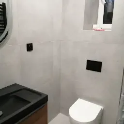 Nowoczesna łazienka z szarymi płytkami, podwieszaną toaletą, czarną umywalką na drewnianej szafce i oknem z widokiem na zewnątrz.