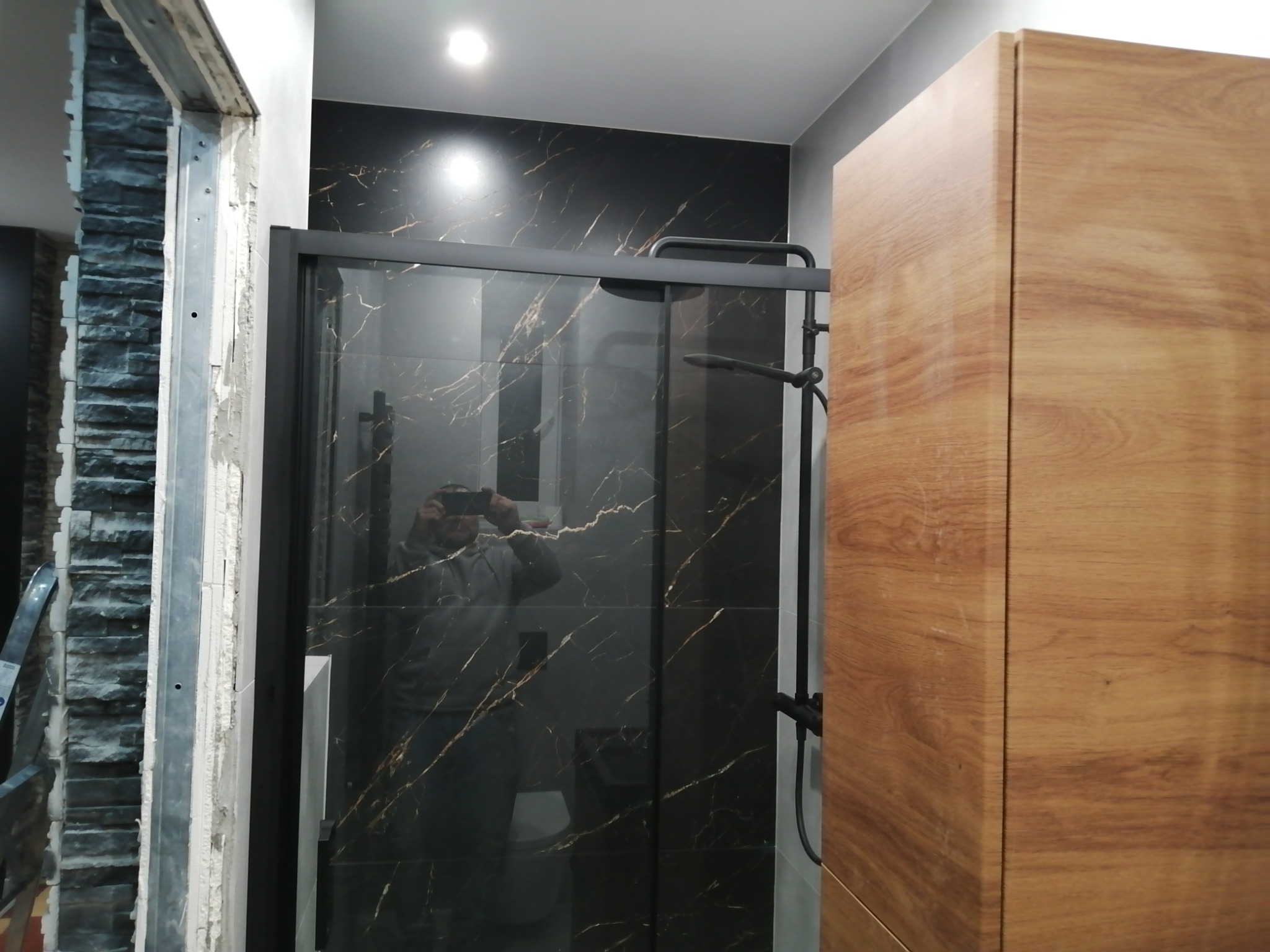 Częściowo wykończona łazienka z czarnymi płytkami imitującymi marmur, kabiną prysznicową z czarnymi profilami i drewnianą szafką. Widoczny fragment ściany z surową cegłą i fragment drabiny.