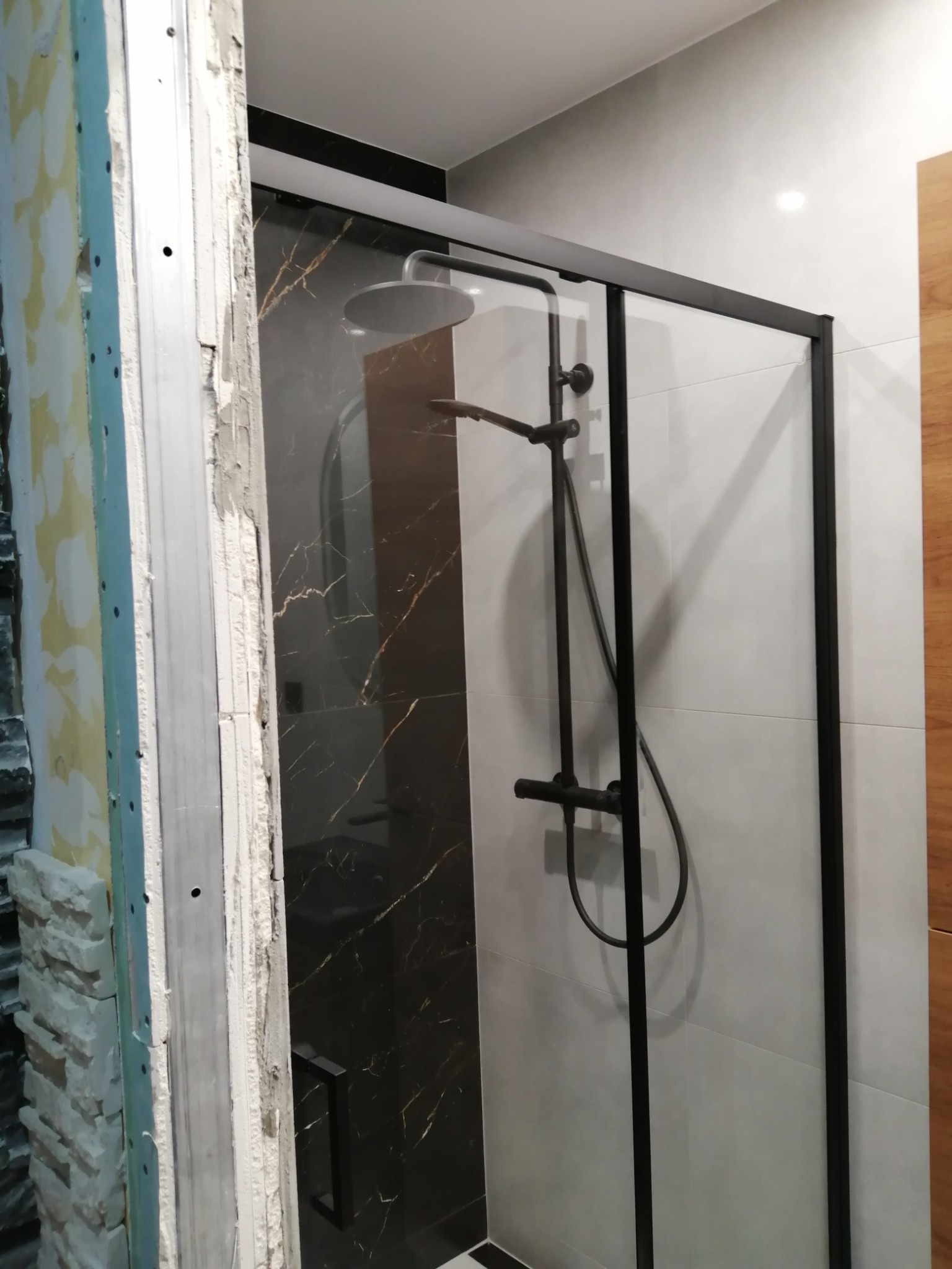 Montaż kabiny prysznicowej z czarną ramą i deszczownicą na tle płytek imitujących marmur, widoczny surowy stan ściany z lewej strony.