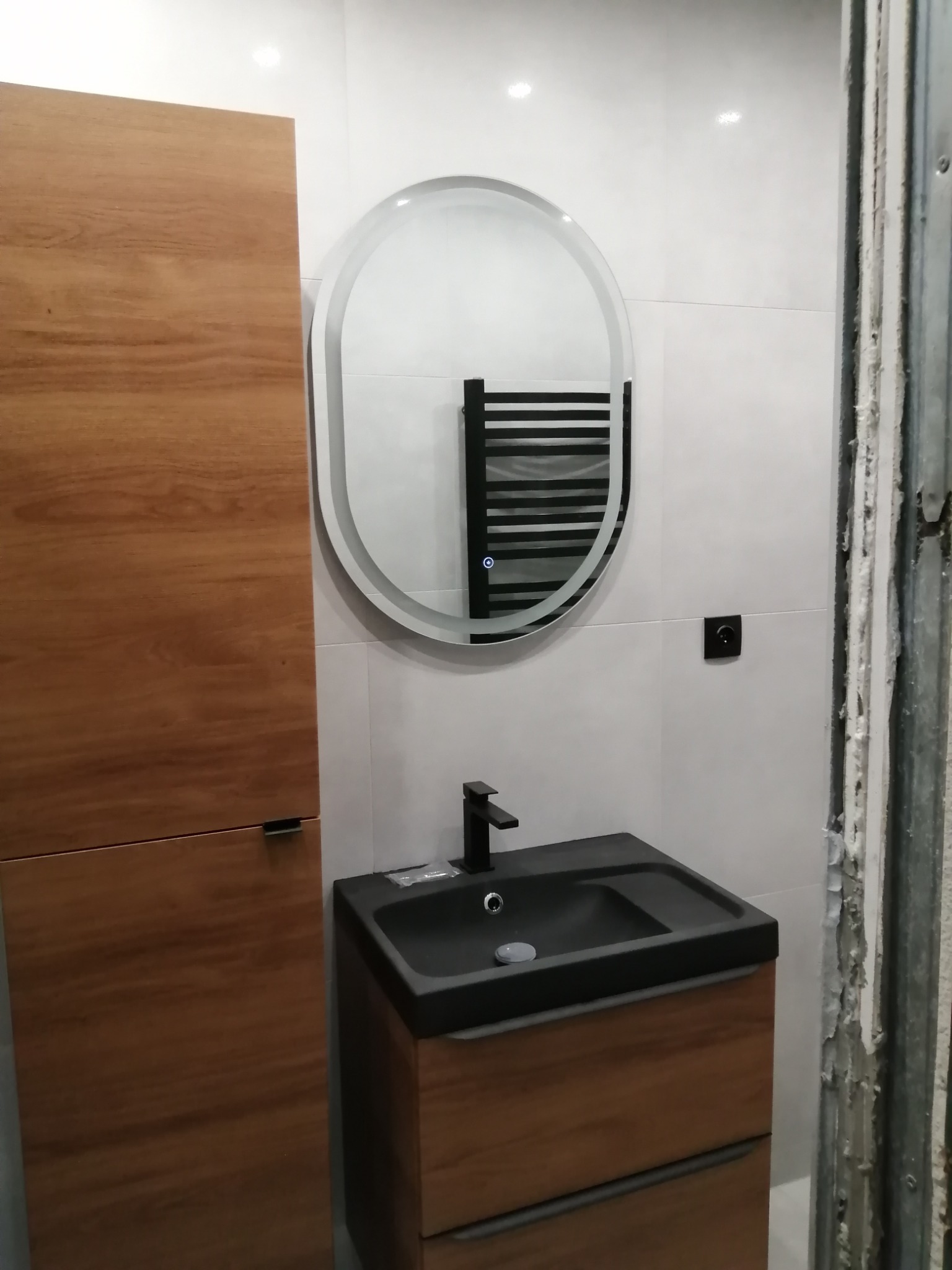 Nowoczesna łazienka z umywalką w kolorze czarnym, szafką z drewnianym fornirem, owalnym lustrem z podświetleniem LED i grzejnikiem drabinkowym w tle, widok na niedokończone wykończenie ściany po...