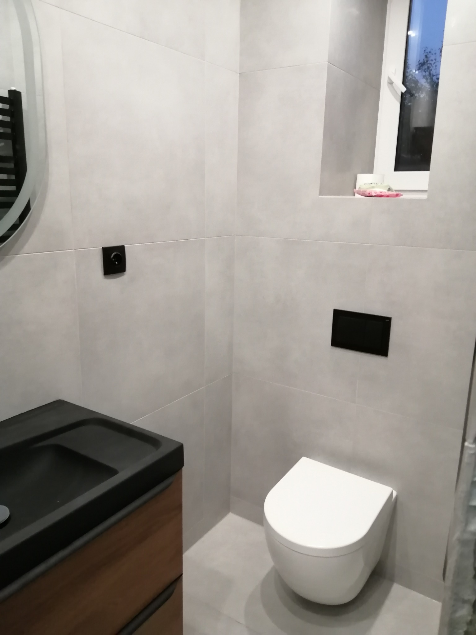 Nowoczesna łazienka z szarymi płytkami, podwieszaną toaletą, czarną umywalką na drewnianej szafce i oknem z widokiem na zewnątrz.