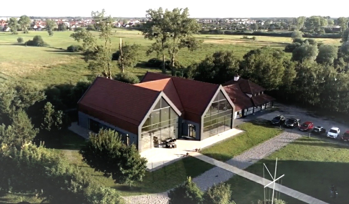 Nowoczesny budynek usługowy z dużymi przeszkleniami, czerwonym dachem i parkingiem, otoczony zielenią, widok z lotu ptaka.