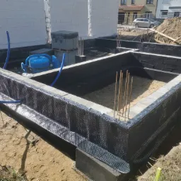 Fundamenty domu w trakcie budowy: hydroizolacja pionowa z folii kubełkowej, wystające pręty zbrojeniowe, widoczna betoniarka, w tle budynek mieszkalny.