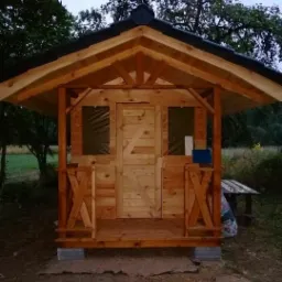 Drewniana altana z zadaszeniem, widoczne detale konstrukcyjne, balustrada z motywem krzyża, drzwi z desek, całość posadowiona na betonowych bloczkach, w tle zieleń.