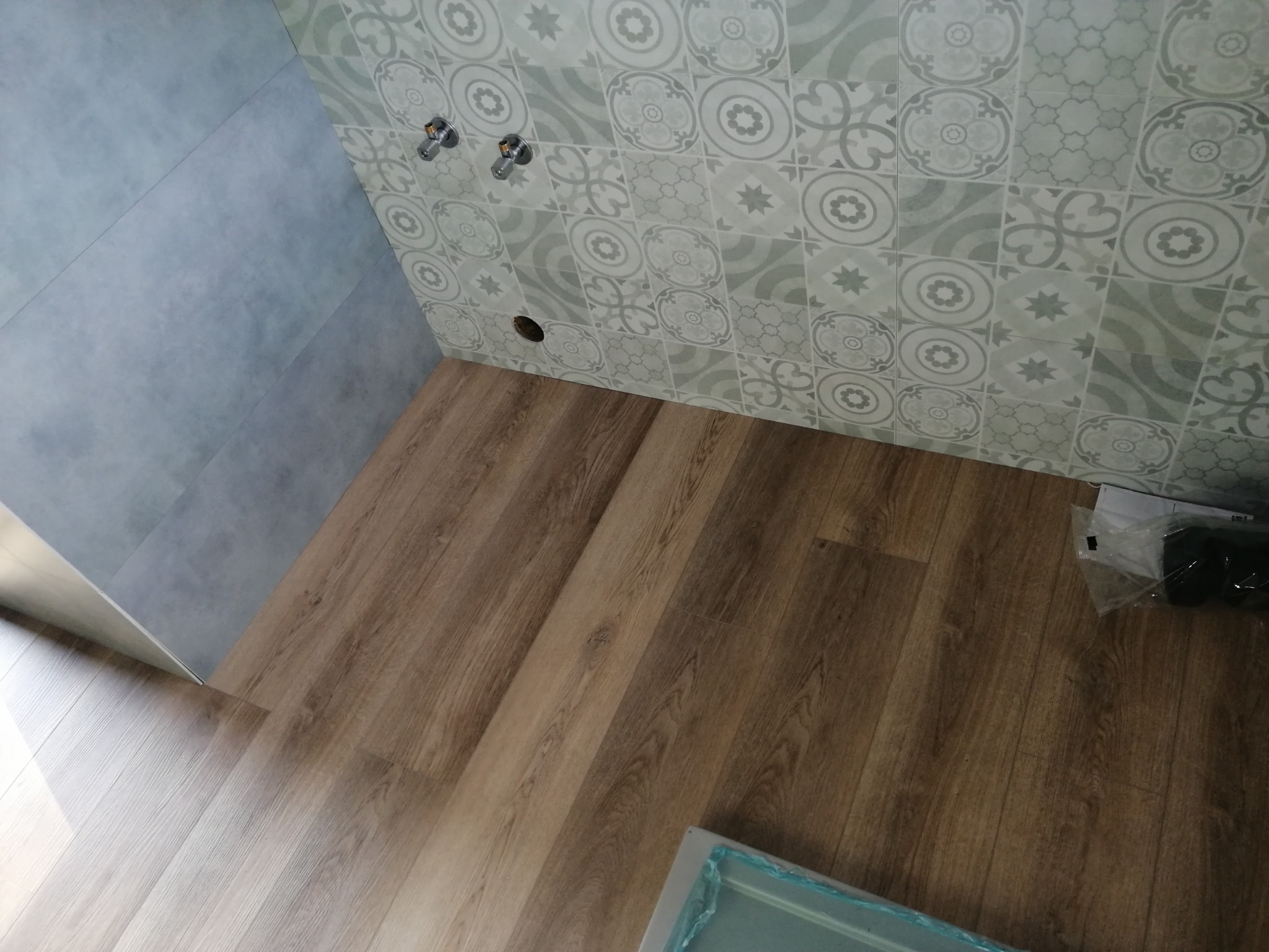 Fragment łazienki w trakcie prac: ułożona podłoga z paneli imitujących drewno, ściana wyłożona szarymi płytkami oraz ściana z dekoracyjnymi płytkami w geometryczne wzory, widoczne przyłącza wodne.