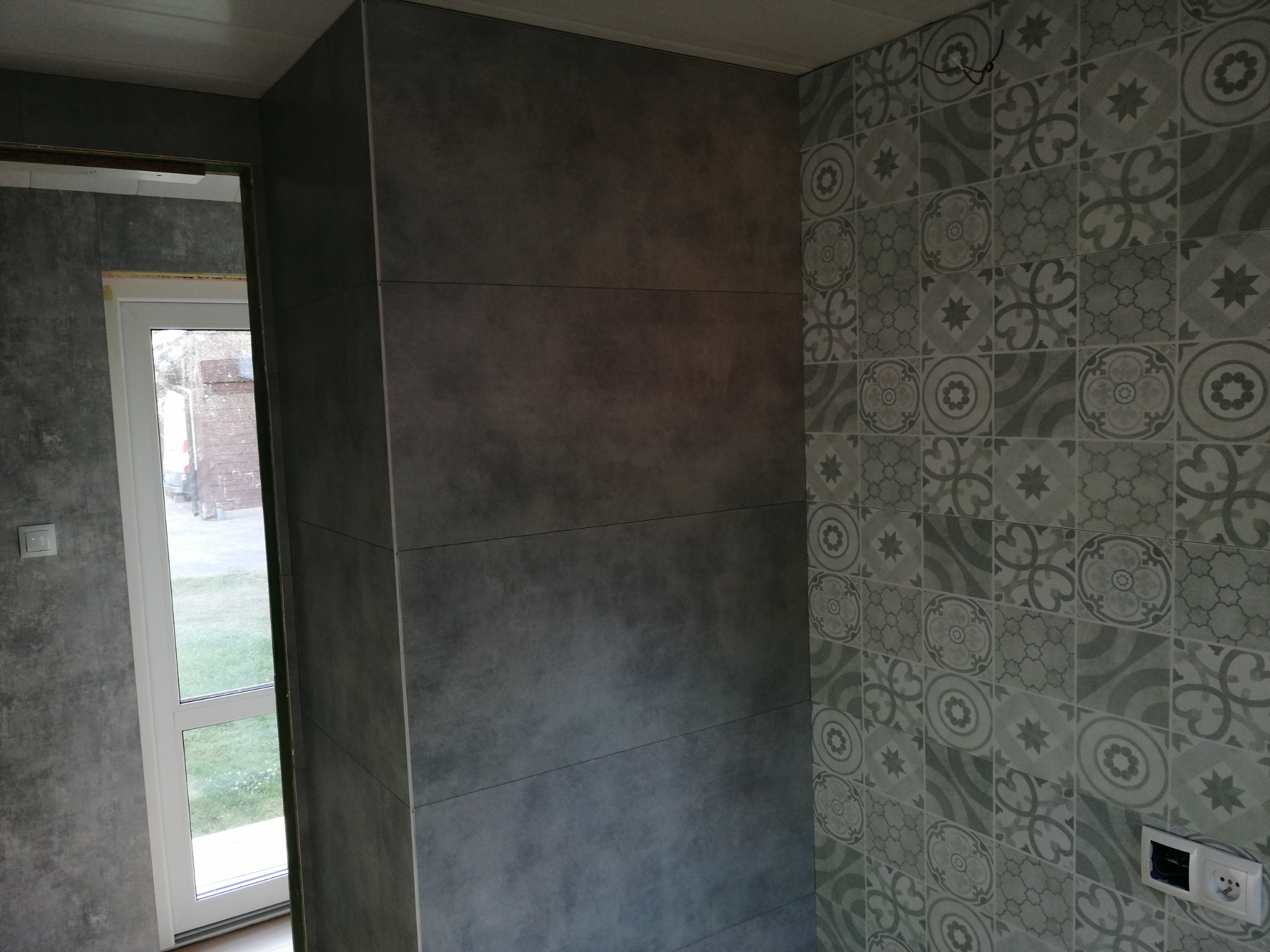 Wykończona ściana w łazience z szarymi płytkami imitującymi beton i dekoracyjnymi płytkami w odcieniach szarości i zieleni, widoczne gniazdko elektryczne i okno.