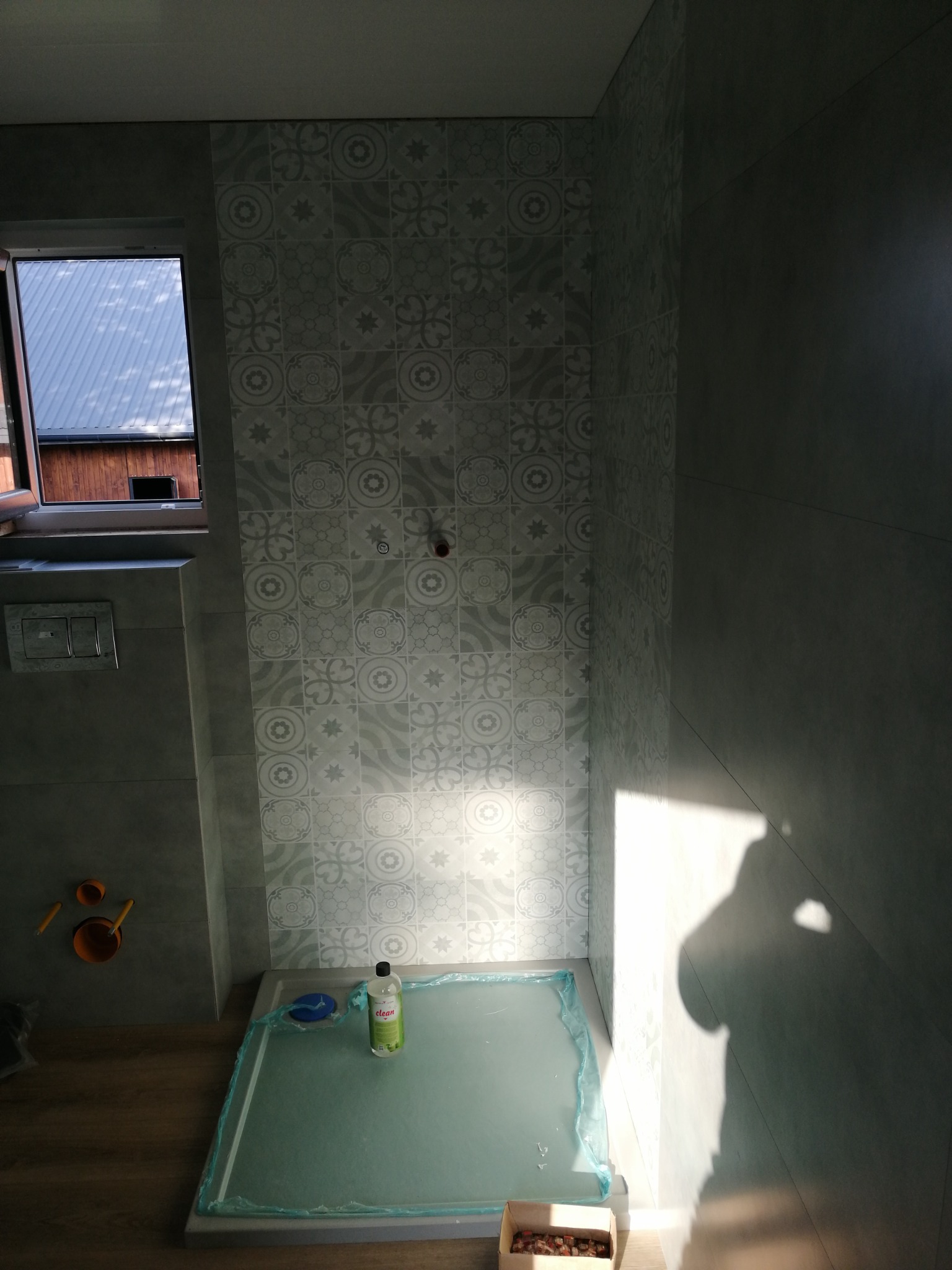 Montaż brodzika w łazience z dekoracyjnymi płytkami ściennymi, widoczne akcesoria instalacyjne i otwarte okno z widokiem na dach budynku.