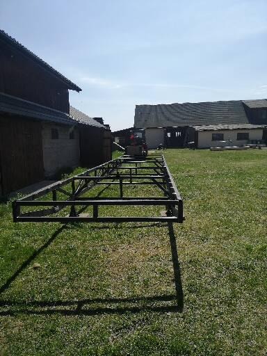 Metalowa, spawana konstrukcja szkieletu domu, ustawiona na trawie w słoneczny dzień, z widocznymi budynkami gospodarczymi i traktorem w tle.