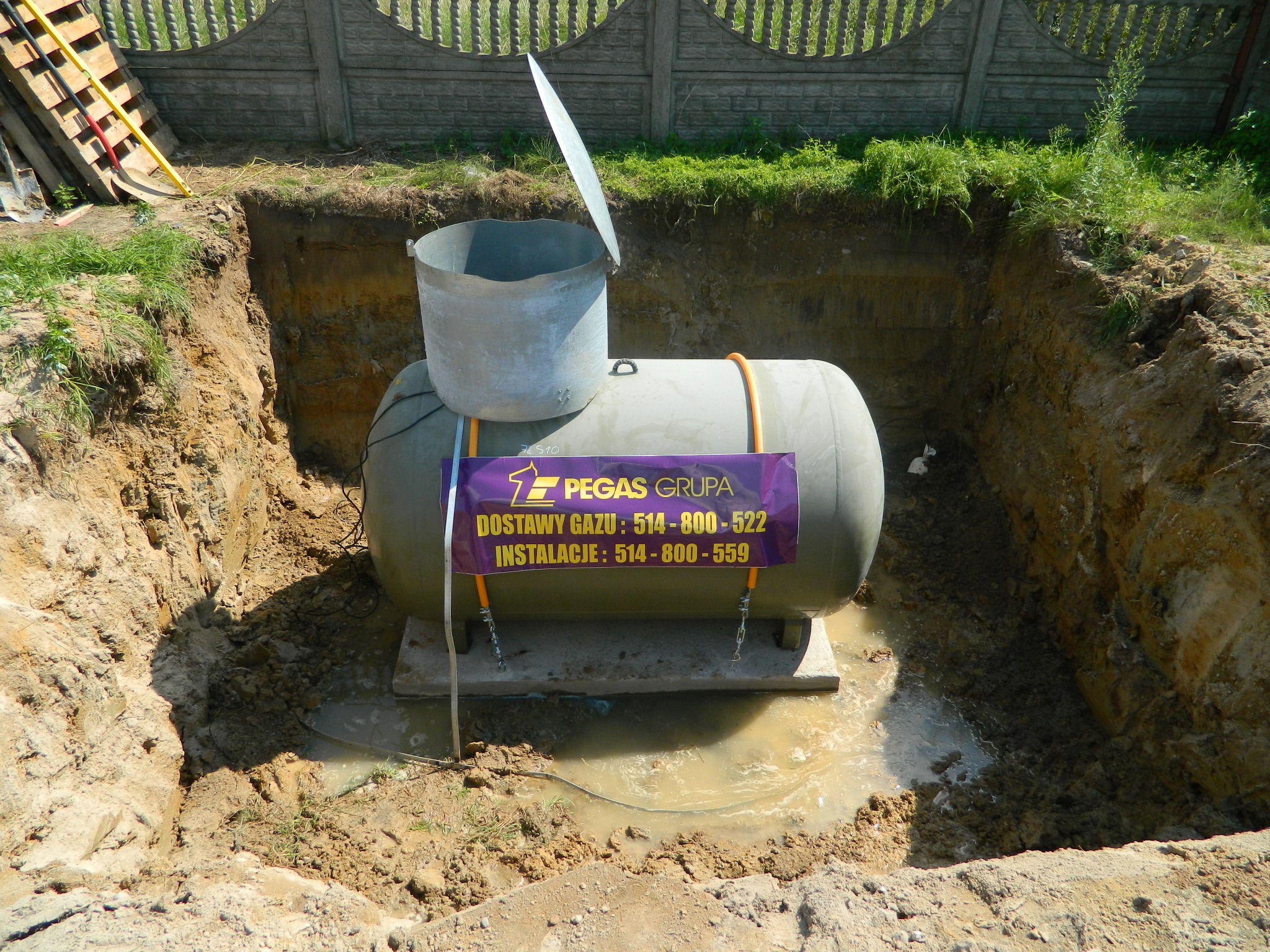 Podziemny zbiornik z metalową osłoną w wykopie, widoczna tabliczka z numerami telefonów firmy Pegas Grupa, woda w wykopie.