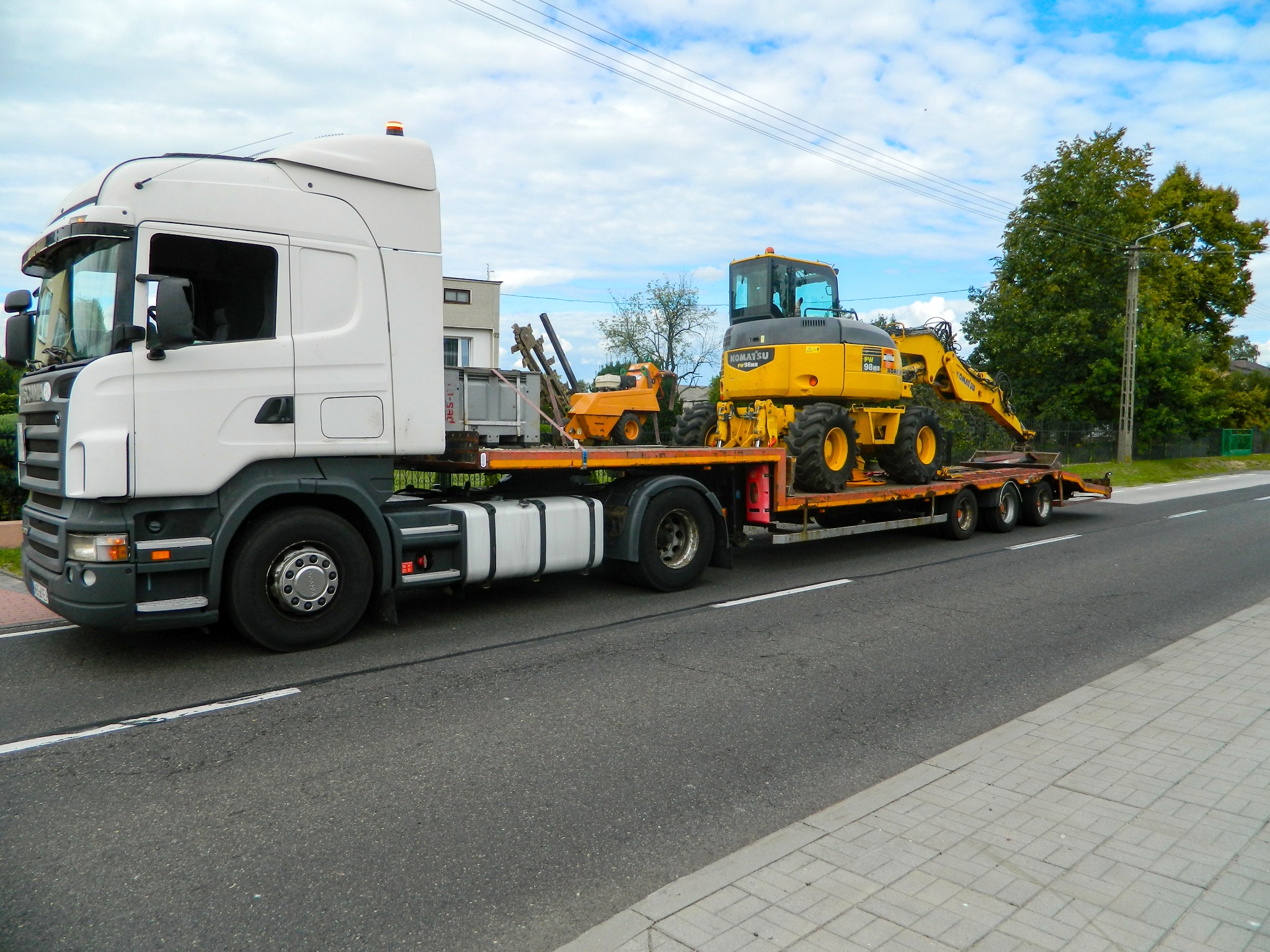 Ciężarówka transportuje dwie żółte maszyny budowlane, w tym koparkę Komatsu, na niskopodwoziowej platformie po asfaltowej drodze w słoneczny dzień.