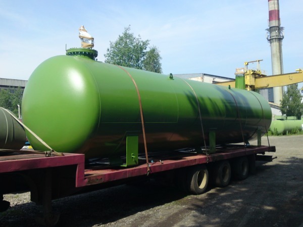 Zielony, cylindryczny zbiornik na gaz przewożony na platformie ciężarowej, z kominem fabrycznym w tle.