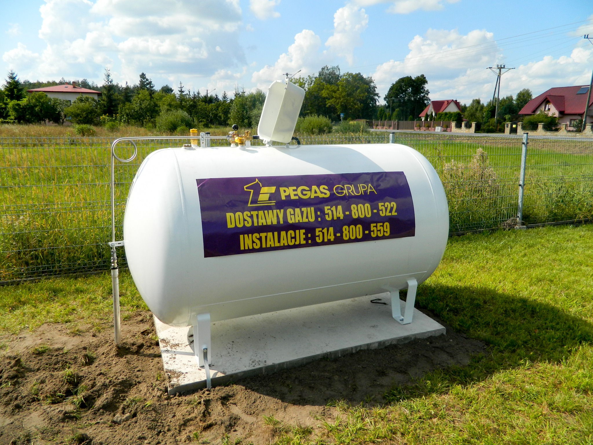 Biały zbiornik na gaz naziemny z otwartą pokrywą, umieszczony na betonowej podstawie na trawniku. Na zbiorniku fioletowa naklejka z logo i numerami telefonów.