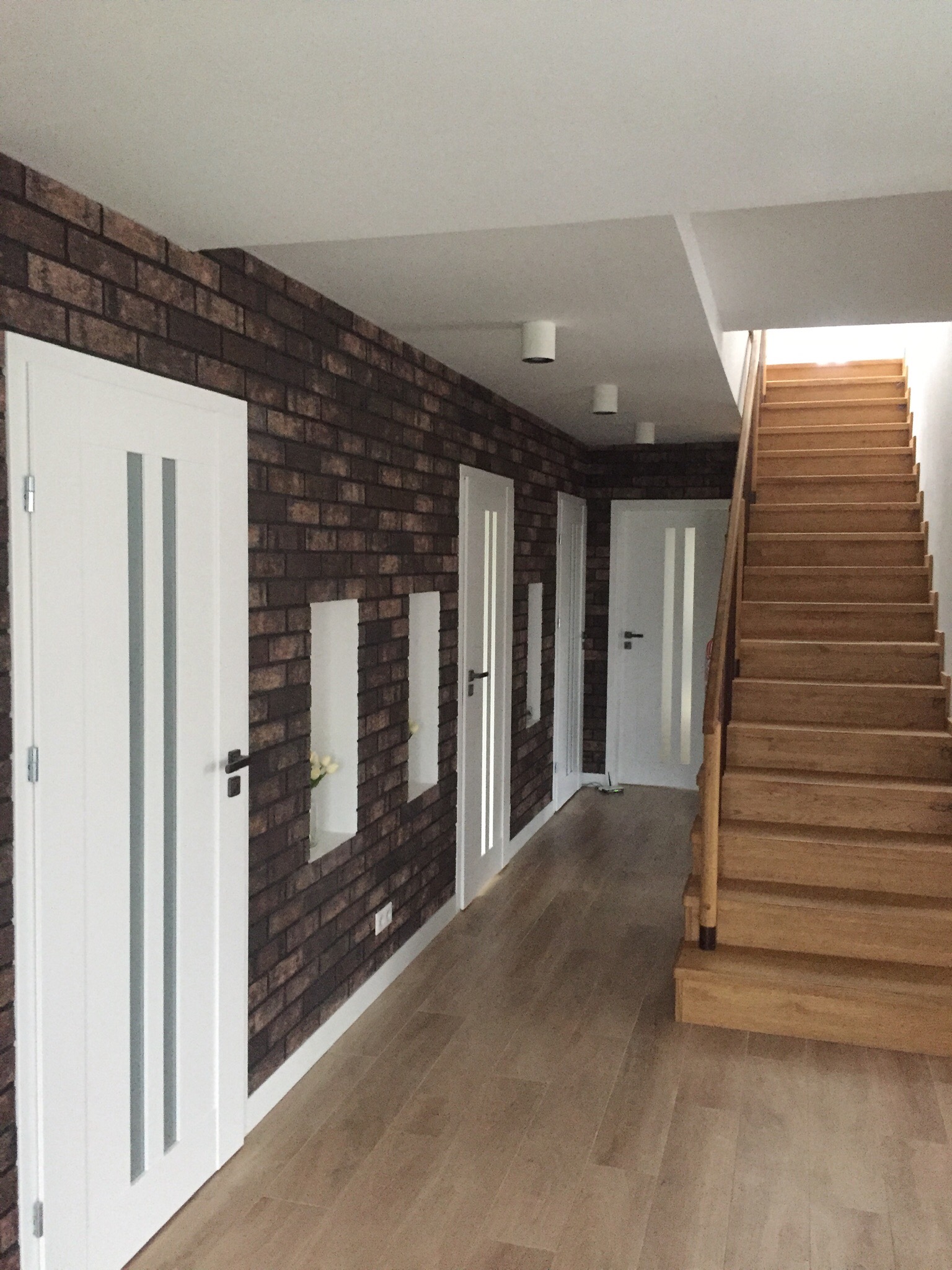 Korytarz z drewnianymi schodami, podłogą imitującą drewno i ścianą wyłożoną tapetą imitującą cegłę, z wnękami dekoracyjnymi i białymi drzwiami z przeszkleniami.