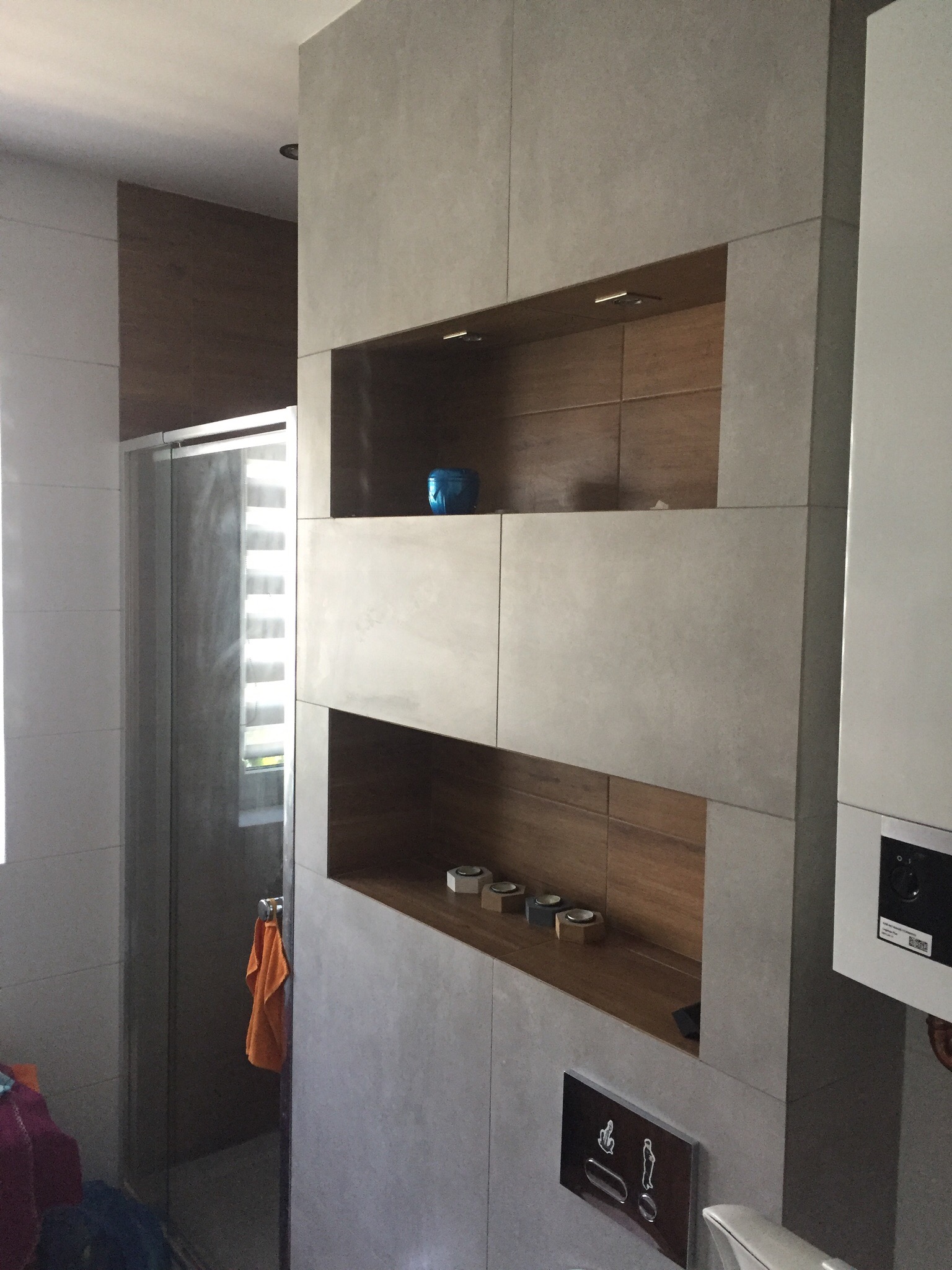 Wykończona łazienka w Kolbuszowej z szarą płytką imitującą beton, zabudową z półkami z drewnopodobną okładziną oraz kabiną prysznicową z widoczną roletą.