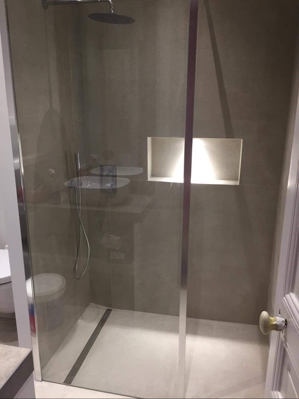 Nowoczesna kabina prysznicowa z szarymi płytkami, podświetloną wnęką i odpływem liniowym w podłodze. Widoczny fragment toalety i drzwi z ceramiczną gałką.