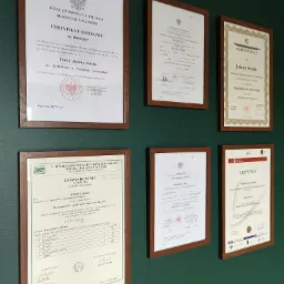 Ściana biura z ramkami zawierającymi certyfikaty i dyplomy Jolanty Soszki, potwierdzające kwalifikacje w dziedzinie księgowości i finansów, zróżnicowane style i formaty dokumentów na zielonym tle...