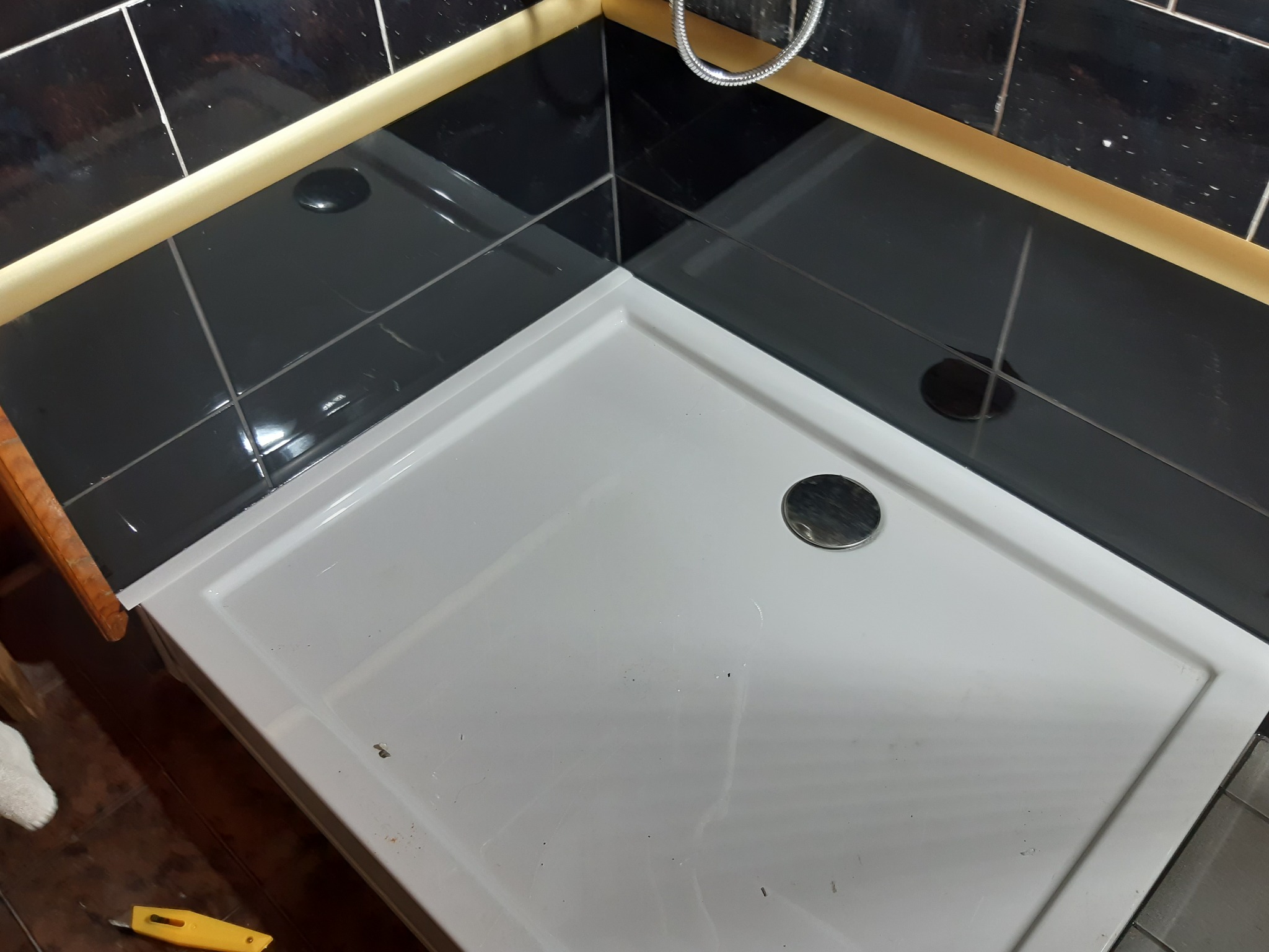 Biały brodzik w rogu łazienki z czarnymi, błyszczącymi płytkami ułożonymi w szachownicę i srebrną słuchawką prysznicową w tle, widoczny fragment ręcznika i żółty nożyk do tapet.