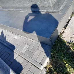 Wykończona podstawa nagrobka z szarych płytek ceramicznych i kostki brukowej ułożonej w jodełkę, cień fotografa z telefonem.