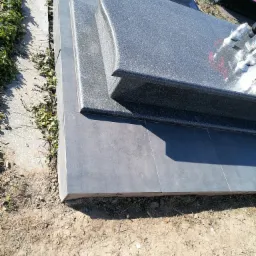 Szary, granitowy nagrobek z lampionami i kwiatami, otoczony płytkami i ziemią.