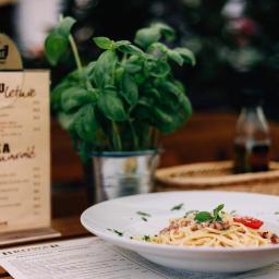 Browar Tarnowski - Kremowe spaghetti carbonara z bazylią i pomidorem na białym talerzu, obok menu z pizzą i piwem Tyskie na drewnianym stole w restauracji.