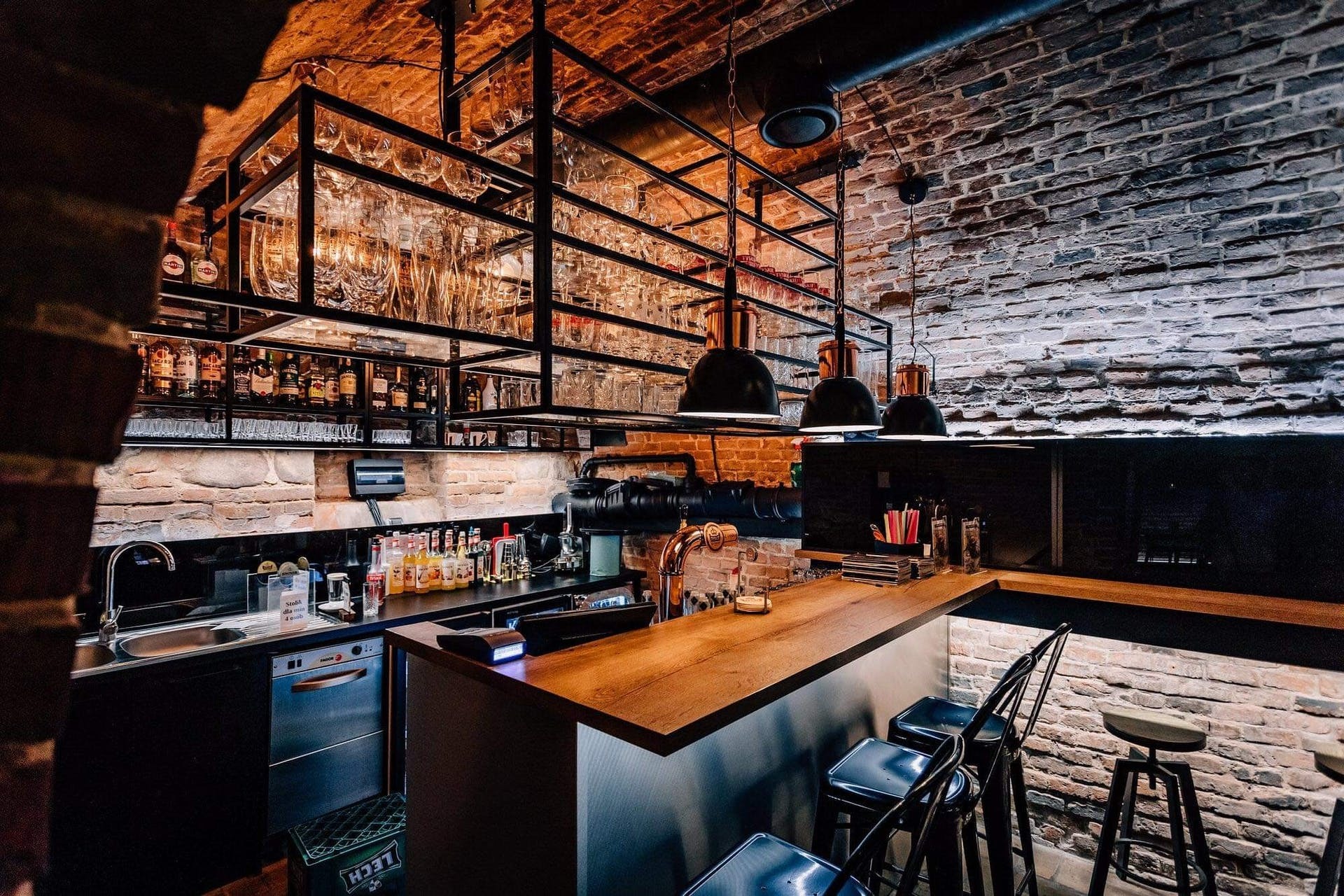 Rustykalny bar z ceglanymi ścianami, oświetleniem w stylu loft i metalowymi półkami z kieliszkami oraz butelkami alkoholi