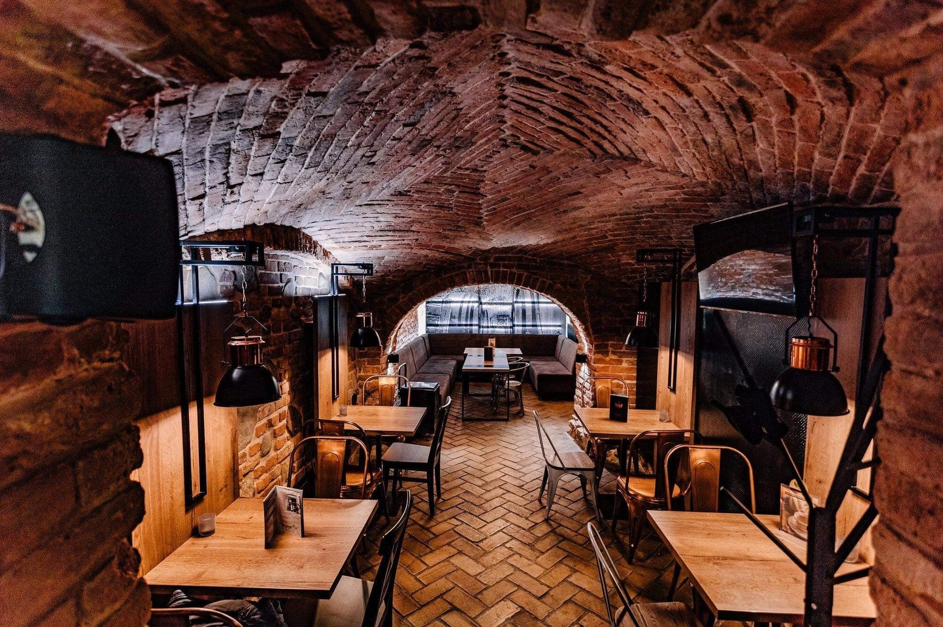 Klimatyczne wnętrze restauracji z ceglanym sklepieniem łukowym, drewnianymi stołami i metalowymi krzesłami, oświetlone wiszącymi lampami w stylu industrialnym.