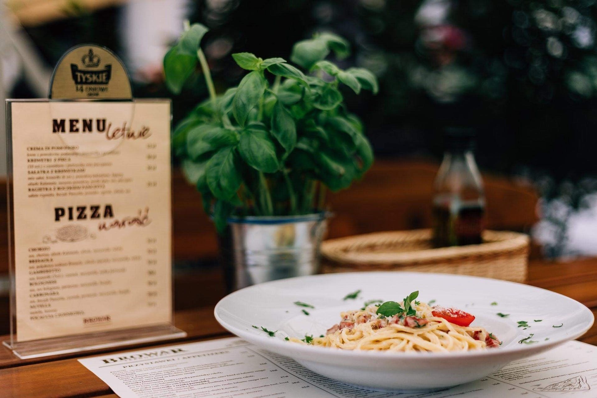 Kremowe spaghetti carbonara z bazylią i pomidorem na białym talerzu, obok menu z pizzą i piwem Tyskie na drewnianym stole w restauracji.
