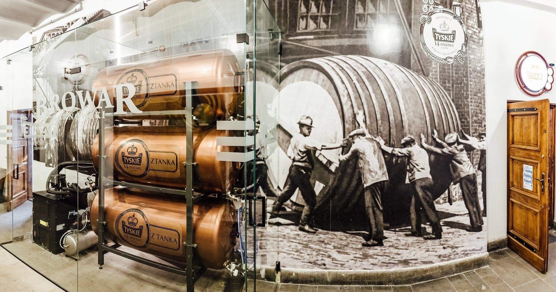 Wnętrze browaru z miedzianymi zbiornikami na piwo 'Tyskie z tanka' za szklaną ścianą, na której widoczna jest grafika przedstawiająca historyczną scenę transportu beczki piwa.