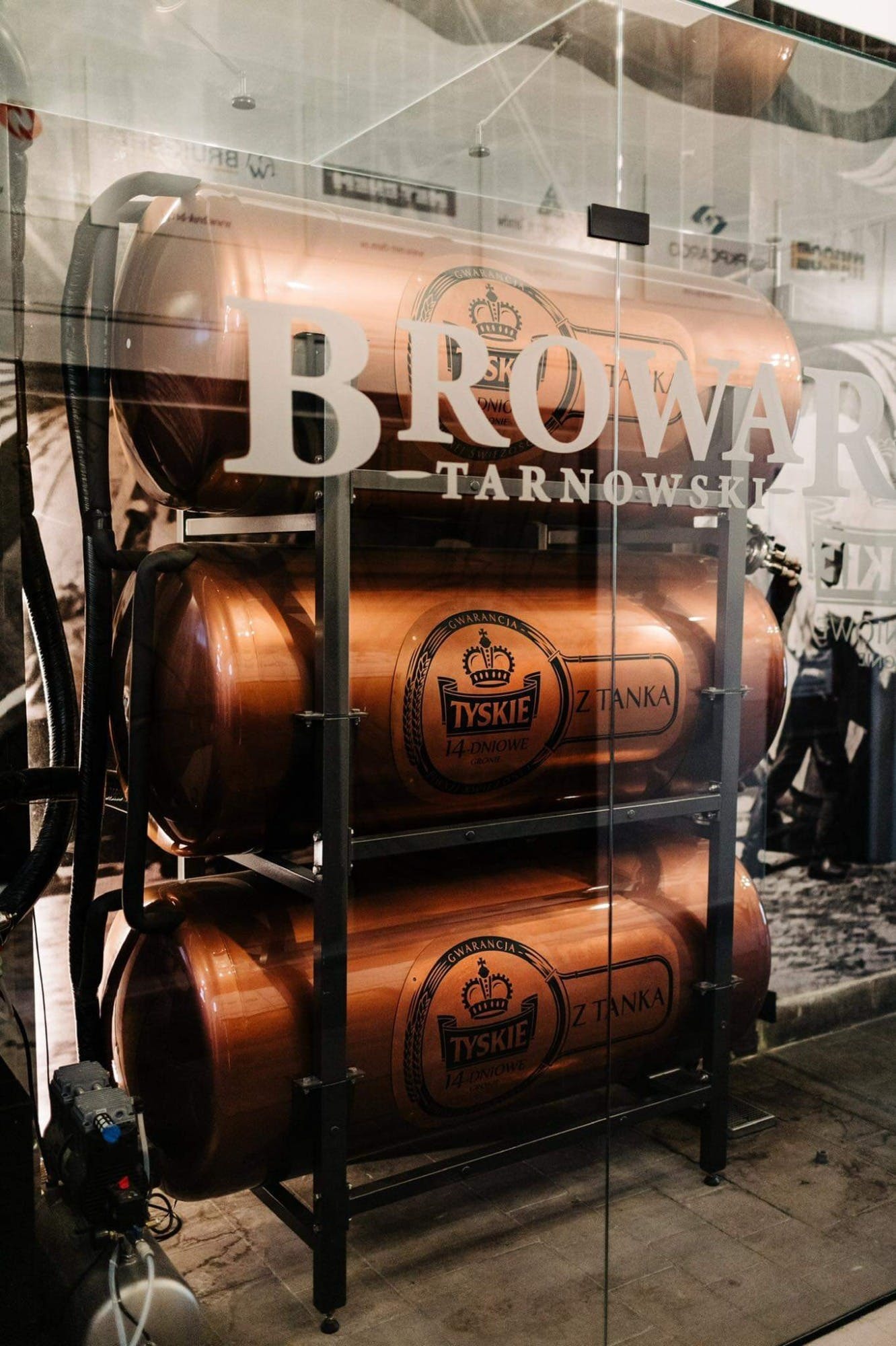 Miedziane zbiorniki browaru Tyskie ustawione jeden na drugim, widoczne przez szybę z napisem Browar Tarnowski.